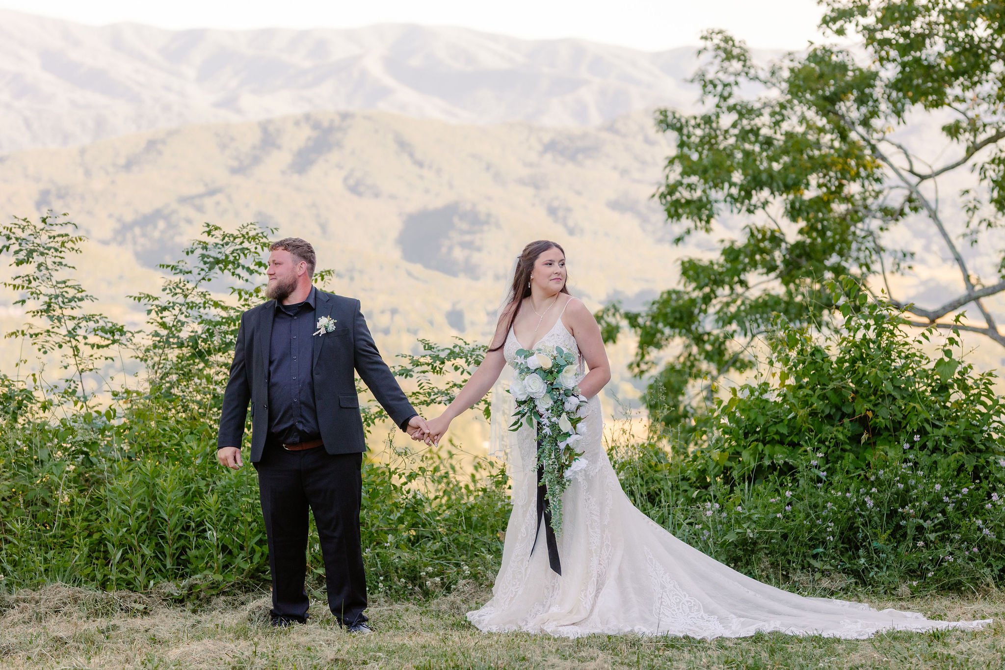 Bride and groom hold hands at a scenic Smoky Mountain overlook, with her lace wedding gown and cascading bouquet framed by mountain ridges during their foothills parkway elopement.