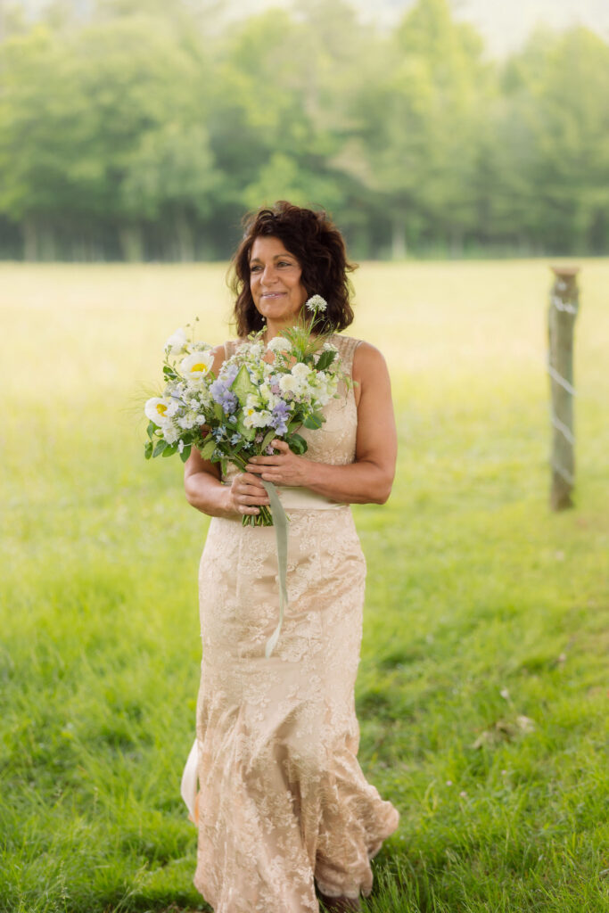 Bride in a champagne floral lace gown holds a loose bouquet of white and lavender wildflowers while standing in a sunlit meadow during her cades cove elopement.