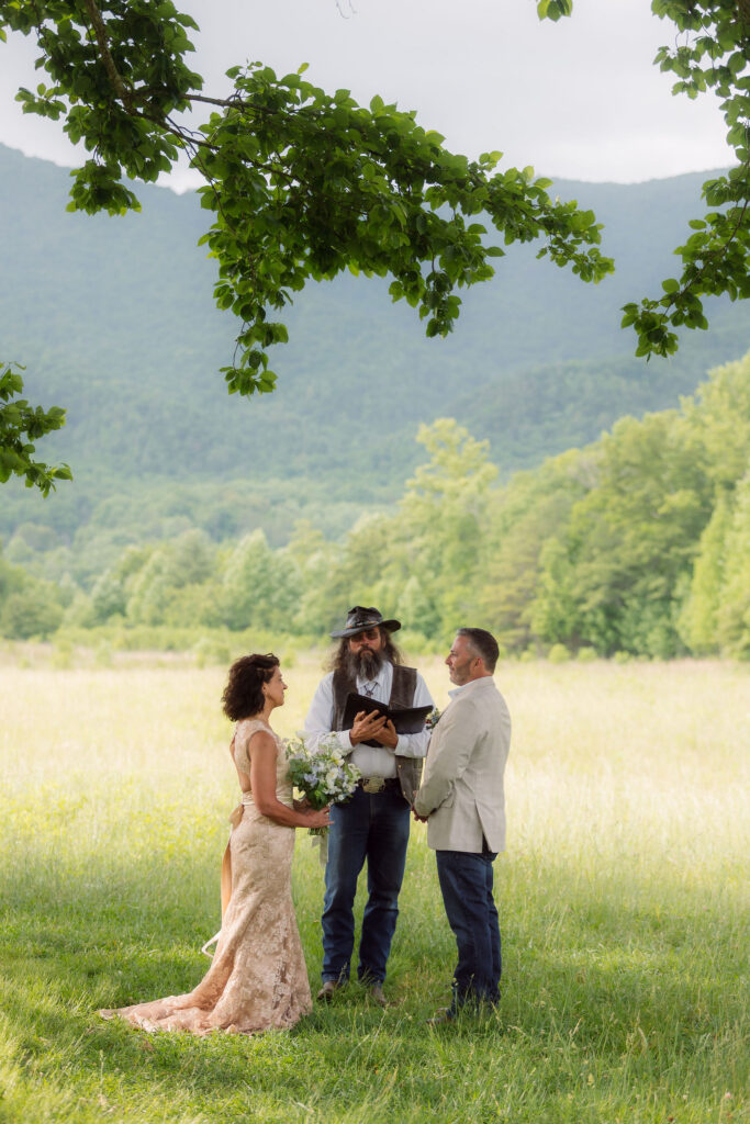 Bride and groom exchange vows beneath a leafy tree as their officiant leads an intimate cades cove elopement ceremony, surrounded by open meadows and the Smoky Mountains.