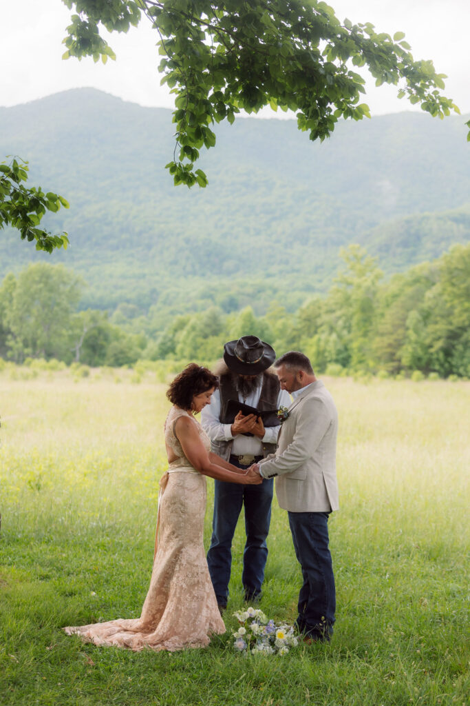 Couple holds hands and bows their heads as their officiant reads from a book beneath leafy branches during an intimate smoky mountain elopement ceremony.