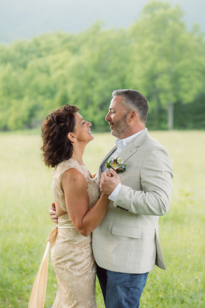 Newlyweds hold hands and smile at each other during their cades cove elopement, with the bride in a champagne lace gown and groom in a beige blazer surrounded by a sunlit meadow.