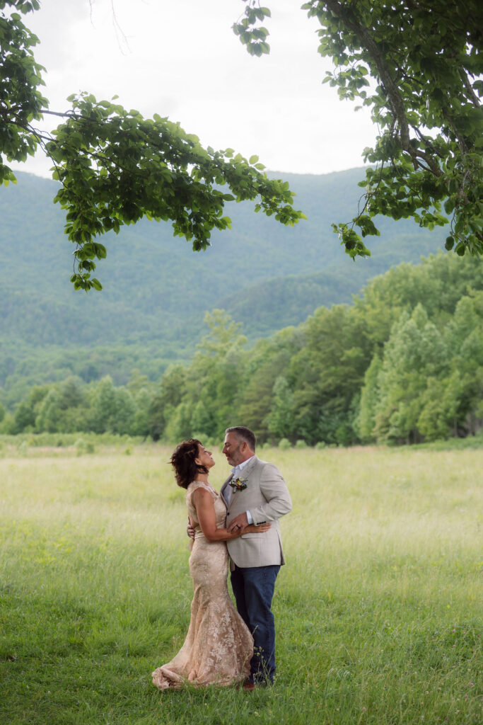 Bride in a champagne lace gown and groom in a light beige jacket embrace beneath leafy trees in a grassy meadow, with mountain views behind them during their cades cove elopement.