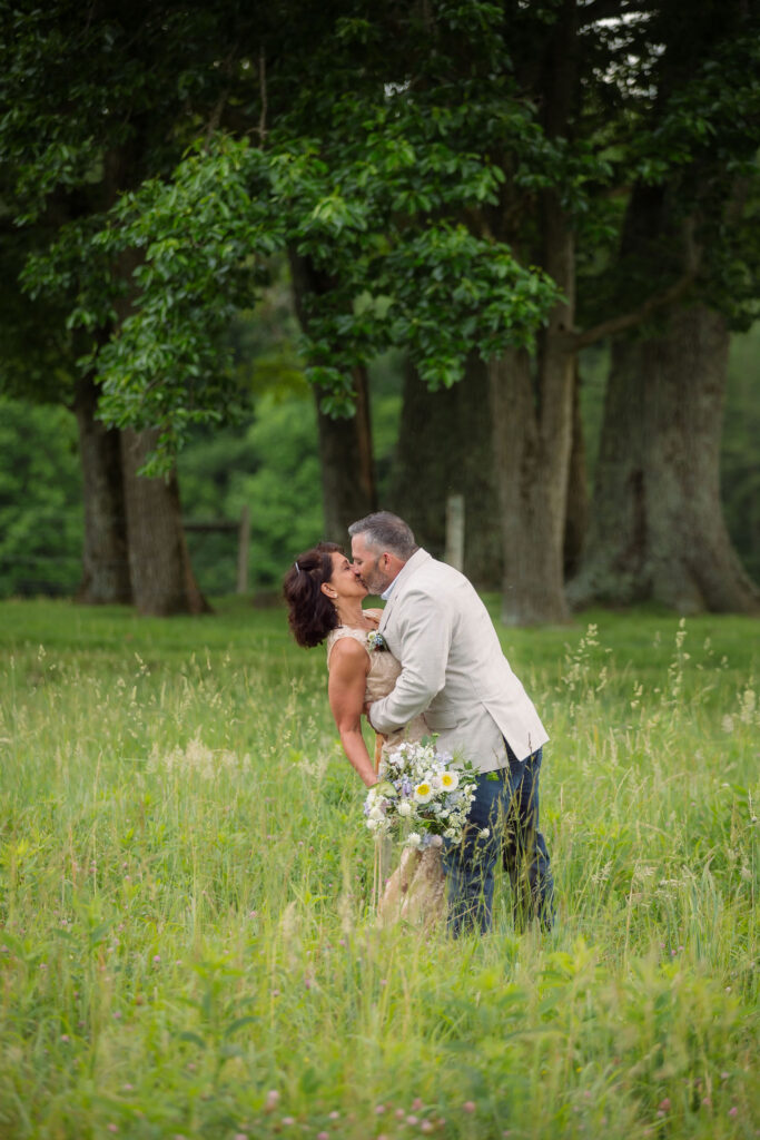 Newlyweds share a kiss in a wildflower-filled meadow beneath towering trees during their smoky mountain elopement in cades cove, with the bride holding a bouquet of white and lavender flowers.