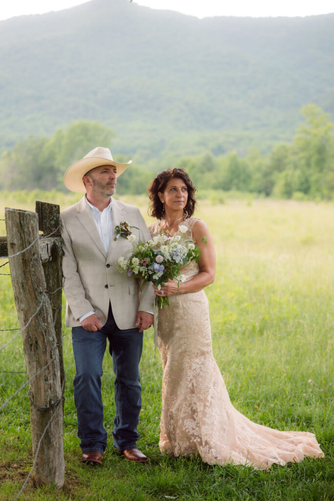 Bride in a champagne lace wedding gown holding a wildflower bouquet beside her groom in a cream cowboy hat and beige jacket, posing by a rustic wooden fence with mountain views during their cades cove elopement.
