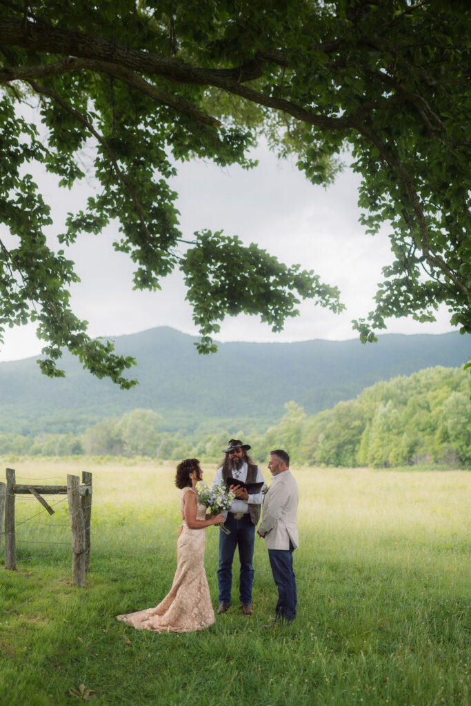 Bride and groom exchange vows beneath a canopy of leafy trees during their cades cove elopement, with a rustic fence, open meadow, and Smoky Mountain peaks framing their outdoor wedding ceremony.