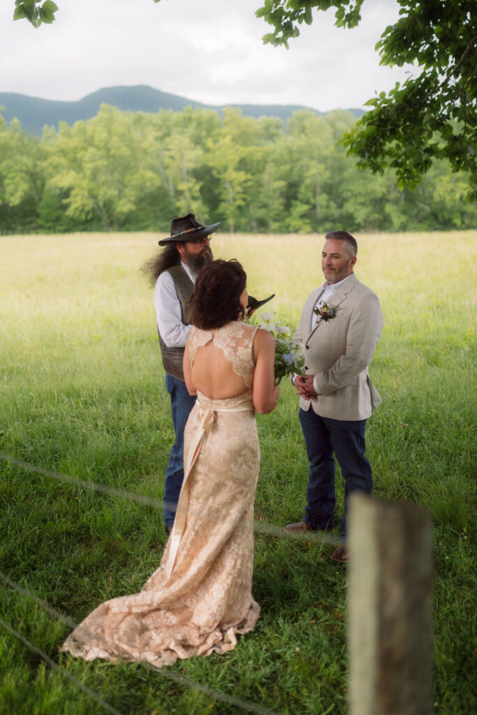 Bride in a backless champagne lace gown faces her groom as he listens during their cades cove elopement ceremony, with a rustic officiant and mountain meadow backdrop.