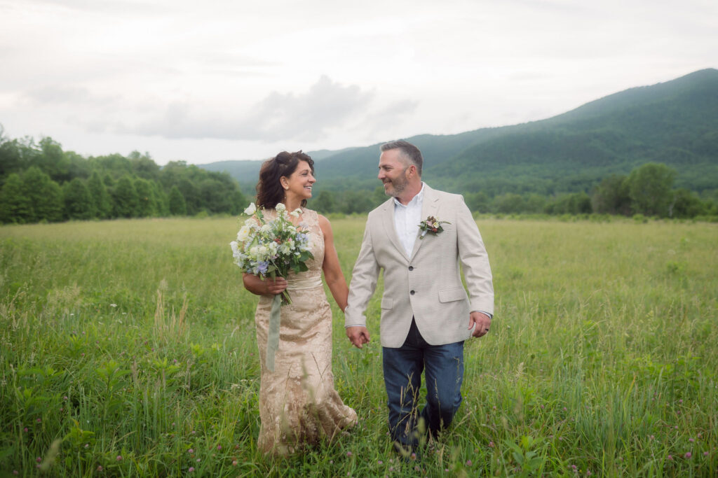Bride in a champagne lace wedding dress and groom in a light beige blazer walk hand in hand through a grassy meadow during their cades cove elopement, with the Smoky Mountains in the background.