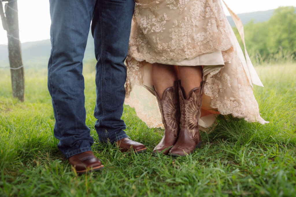 Close-up of the bride’s embroidered brown cowboy boots beneath her champagne lace wedding dress beside the groom’s jeans and leather boots during their cades cove elopement.