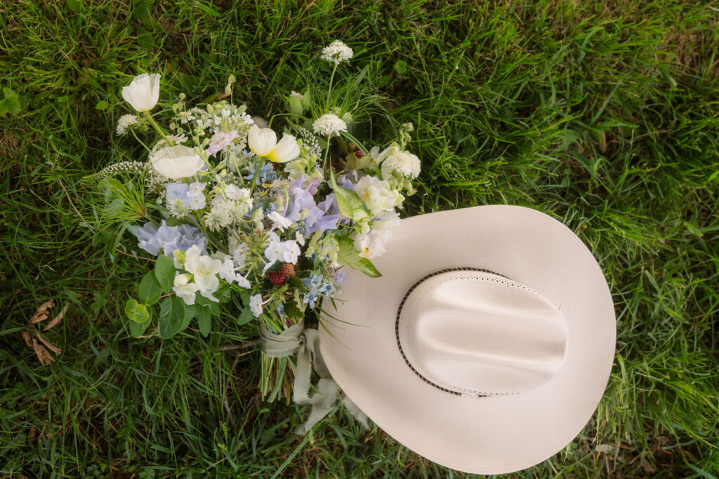 A cream cowboy hat rests beside a wildflower bridal bouquet of white, lavender, and pale blue blooms on green grass during a rustic cades cove elopement.