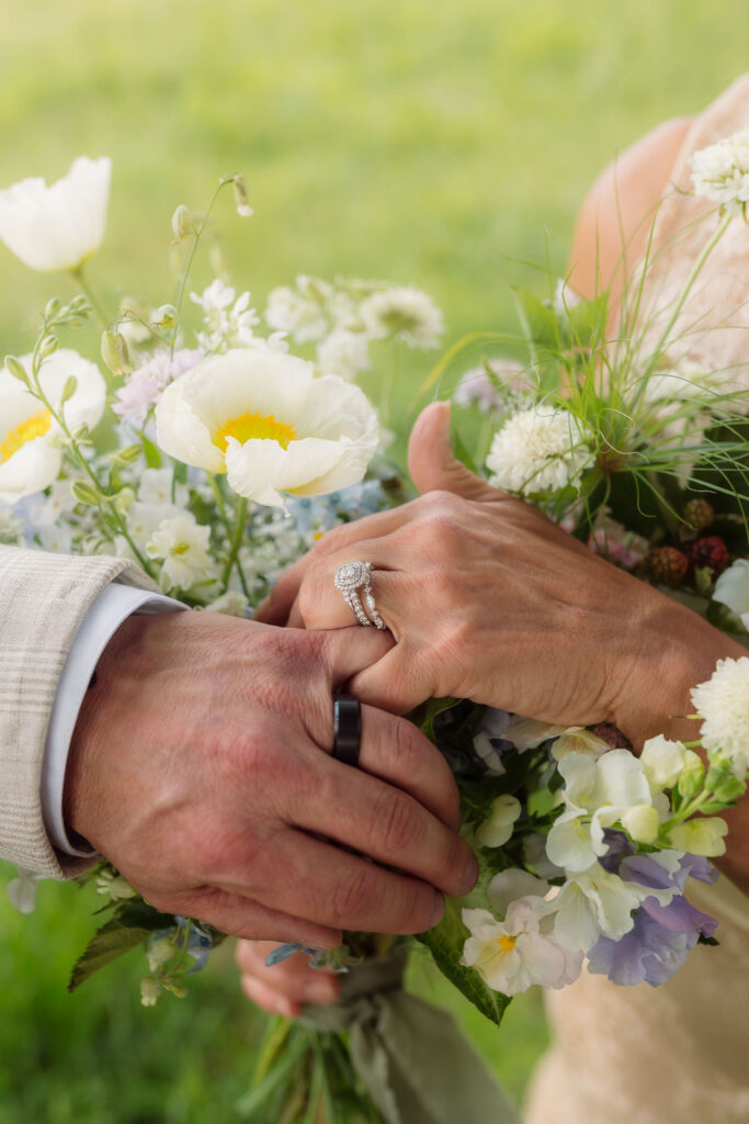 Close-up of a couple’s wedding rings and hands resting on a bouquet of white and lavender wildflowers during their cades cove elopement in the Smoky Mountains.