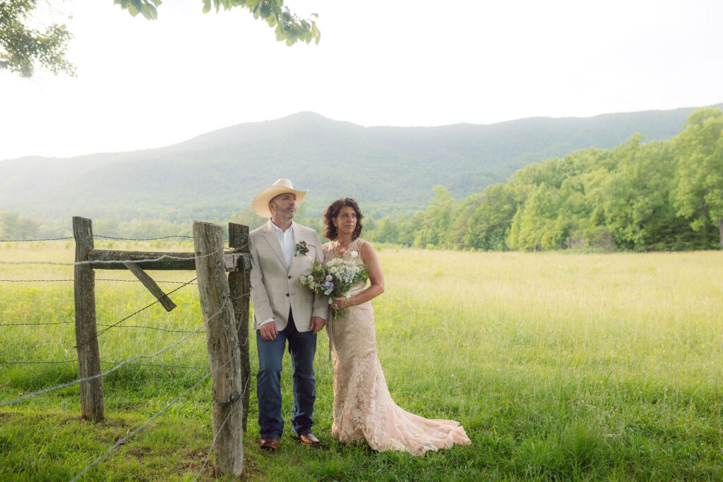 Bride in a champagne lace wedding dress holding a wildflower bouquet beside the groom in a cowboy hat and cream blazer near a rustic wooden fence, with mountain views during their cades cove elopement.