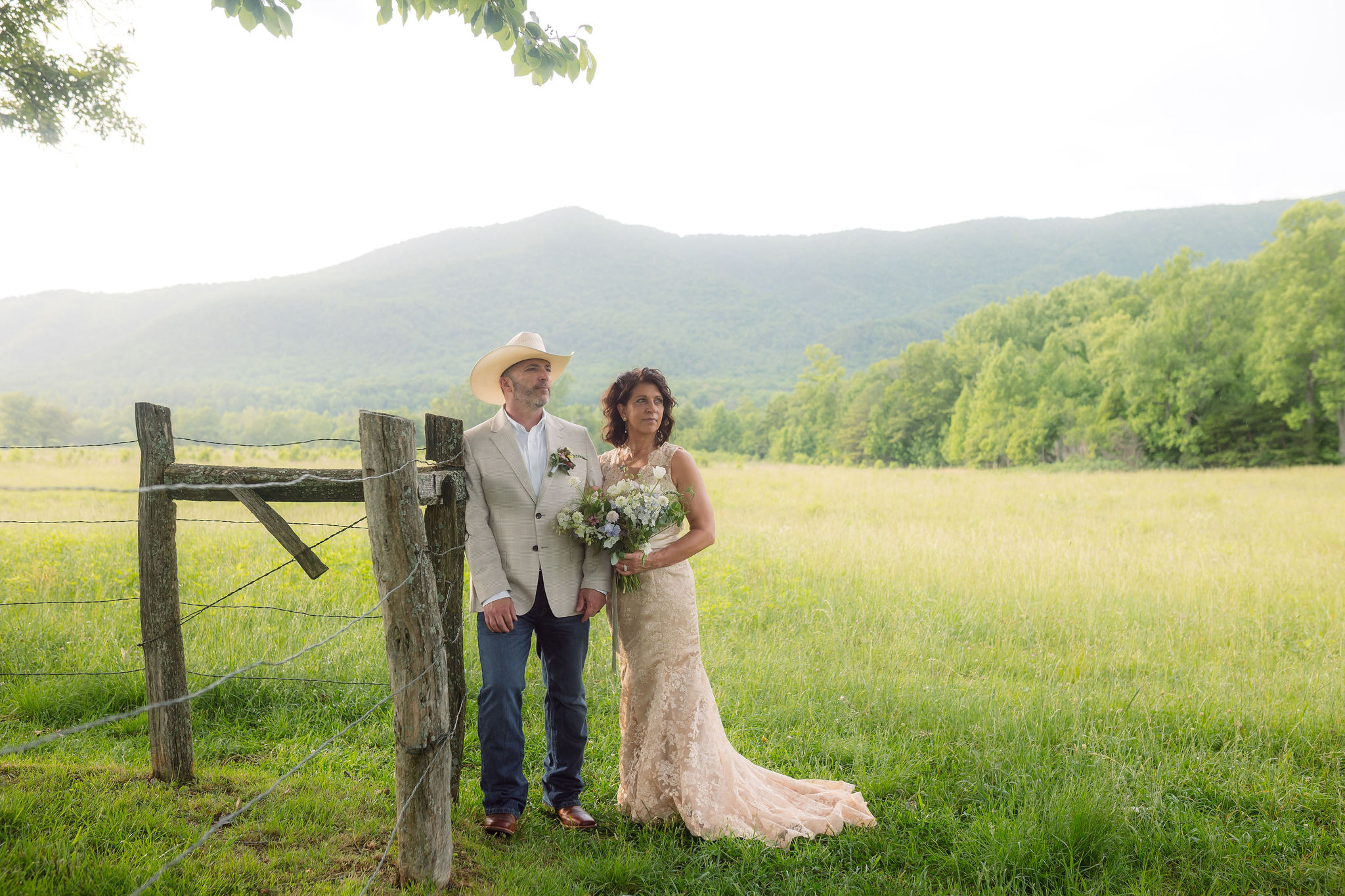 Bride in a champagne lace wedding dress holding a wildflower bouquet beside the groom in a cowboy hat and cream blazer near a rustic wooden fence, with mountain views during their cades cove elopement.